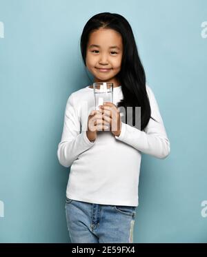 Niedlich lächelnd asiatisch koreanisch Kind Mädchen in Jeans und weiß Langarm-Shirt steht mit vollem Glas reines Wasser Stockfoto