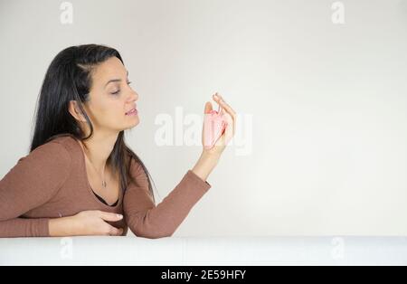 Frau, die ein Herz in der Hand hält und auf dem Sofa vor der weißen Bkank-Wand sitzt. Stockfoto
