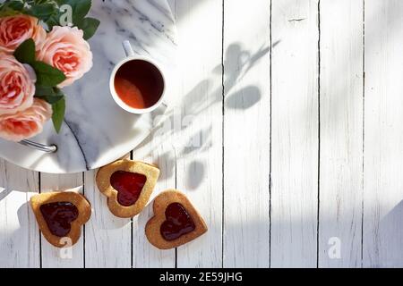 Herzförmige Kekse mit Erdbeermarmelade, Tasse Kaffee, rosa Blumen auf weißem Holztisch. Geburtstag, Valentinstag, Muttertag. Top-Ansicht. Tageslicht Stockfoto