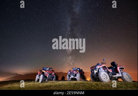 Nachtbild. Drei atv Quad Motorräder stehen auf der Spitze des Berges unter schönem Nachthimmel voller Sterne und Milchstraße im Hintergrund Stockfoto