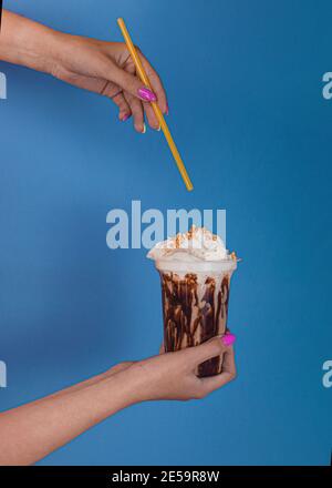 Frau Hand hält ein Glas Milchschokolade Cocktail mit Kaffee mit Schlagsahne auf blauem Hintergrund. Die Hand einer Frau hält einen Cocktailstroh Stockfoto