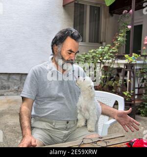 Ein Mann spielt mit seinem kleinen maltesischen Hund. Beide genießen den Schatten der Bäume und den Garten, in dem sie leben. Stockfoto