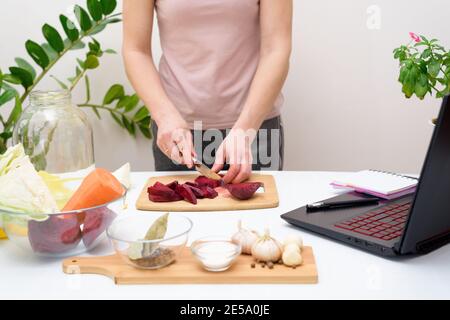 Kochen zu Hause. Frau schaut online Video Rezepte auf einem Laptop und kocht in der Küche zu Hause, Gemüse schneiden. Eingelegter rosafarbener Kohl, fermentiert Stockfoto