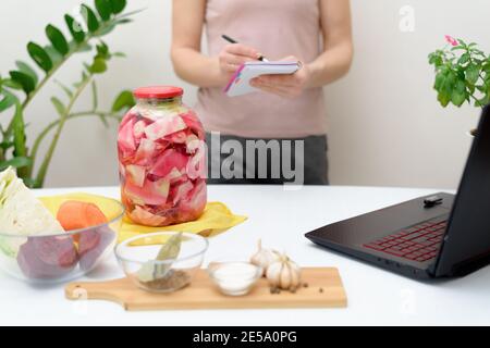 Kochen zu Hause. Eine Frau schaut online Video Rezepte auf einem Laptop und kocht in der Küche zu Hause schreibt in einem Notebook. Eingelegter rosafarbener Kohlfermenta Stockfoto