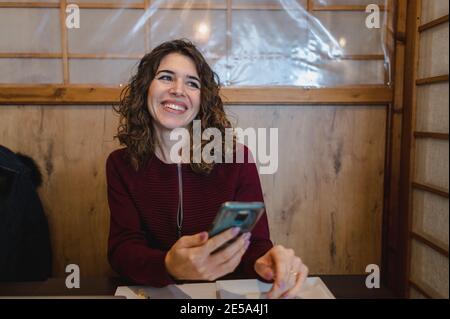 Flacher Fokus einer lockigen Frau, die mit ihrem Smartphone lächelt Ihre Hände Stockfoto