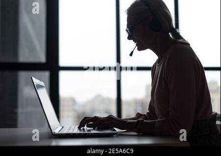 Seitenansicht eine Silhouette einer Callcenter-Mitarbeiterin, die ein Headset und einen Laptop für die Online-Kommunikation mit den Kunden verwendet, wobei eine Frau spricht Stockfoto