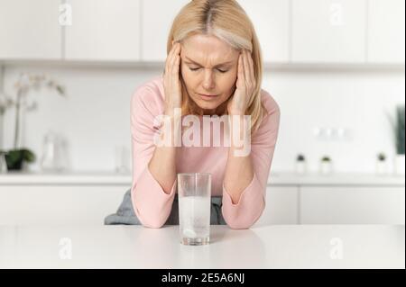 Eine Frau mittleren Alters leidet unter Kopfschmerzen, hält den Kopf, massiert die Schläfen und fühlt sich verletzt, Brauseschmerzlindertablette löst sich im Glas Stockfoto