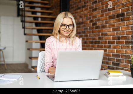 Eine Frau mittleren Alters sitzt auf dem Schreibtisch im Loft-Stil Büro und chattet online auf dem Laptop. Eine fröhliche Geschäftsfrau trägt eine Brille, die antippt Stockfoto