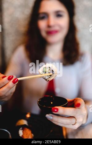 Flacher Fokus einer lächelnden lockigen Frau, die Sushi mit hält Essstäbchen Stockfoto