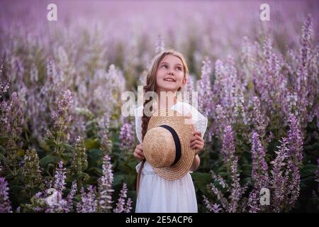 Portraits schöne Teenager-Mädchen in einem Strohhut, mit einem langen Zopf in einem blühenden rosa Salbei Feld. Weiße Sundress. Blumenhintergrund im Sonnenuntergang Licht. R Stockfoto