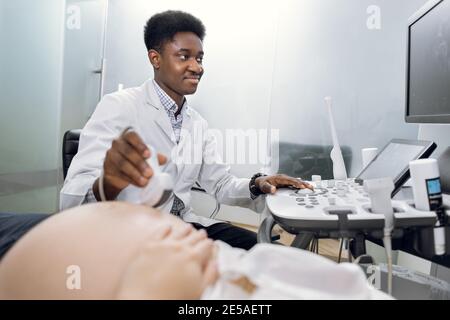 Seitenansicht der angenehmen lächelnden afroamerikanischen männlichen Arzt tun Ultraschall-Scan für unkenntlich schwangere Frau im Krankenhaus. Ultraschalldiagnostik Stockfoto