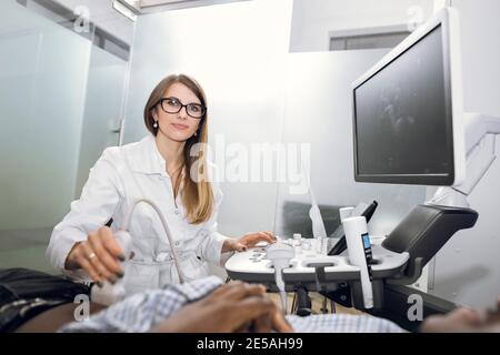 Professionelle Frau Sonographer Therapeut, die mit Ultraschall-Maschine in der Klinik, machen Abdomen Ultraschall für schwarzen männlichen Patienten, Blick auf Stockfoto