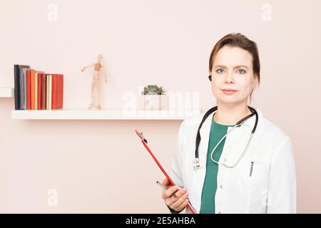 Portrait von selbstbewussten jungen Arzt trägt weißen Mantel mit Stethoskop und Hörmuschel, die in ihrem Home Office stehen und sie betrachten Kamera bereit für onl Stockfoto