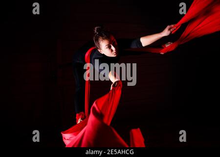 Ein Sportmädchen führt Gymnastik- und Zirkusübungen auf roter Seide durch. Studioaufnahmen vor dunklem Hintergrund. Luftturnen auf Leinwand Stockfoto