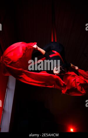 Ein Sportmädchen führt Gymnastik- und Zirkusübungen auf roter Seide durch. Studioaufnahmen vor dunklem Hintergrund. Luftturnen auf Leinwand Stockfoto