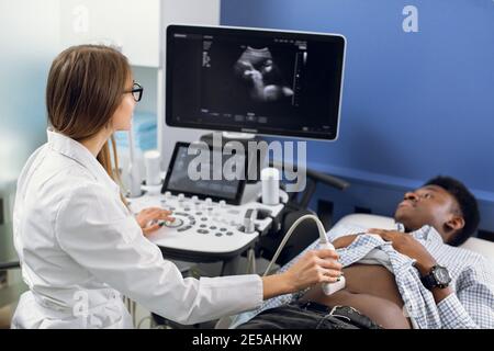 Junge kaukasische Frau Arzt bewegen Ultraschallaufnehmer auf afrikanischen Mannes Bauch im Krankenhaus. Weibliche Arzt macht ultra Klang der inneren Organe von Stockfoto