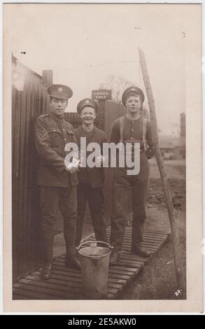 Informelles Foto einer Gruppe von drei Soldaten aus dem Ersten Weltkrieg, die vor einem Schild stehen, auf dem nur die Damen stehen. Einer der Soldaten hat beide Daumen nach oben, ein anderer scheint ein Gemüse zu schälen. Ein Eimer steht auf den Dielen davor. Stockfoto