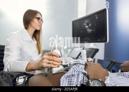 Frau Arzt führt einen Ultraschall des Patienten Bauch und innere Organe, Blick auf den Bildschirm der modernen Ultraschallgerät. Afrikanischer männlicher Patient Stockfoto