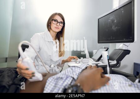 Ultraschall in der Physiotherapie. Junge Frau Therapeut Sonographer, mit Ultraschall-Scanner auf dem Bauch des schwarzen männlichen Patienten. Abdomen und Stockfoto