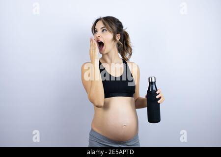 Junge Sportlerin schwanger trägt Sportkleidung mit Flasche mit Wasser über weißem Hintergrund bedeckt Mund mit der Hand, schockiert und Angst vor Fehler Stockfoto