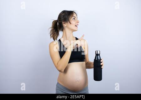 Junge Sportlerin schwanger trägt Sportkleidung hält Flasche mit Wasser über Weißer Hintergrund lächelt und zeigt mit Hand und Finger auf Die Seite Stockfoto