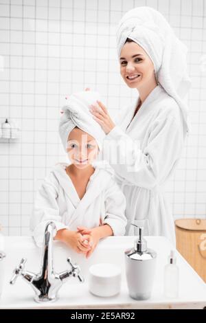 Lächelnde Mutter im Bademantel trägt Handtuch auf dem Kopf des Kindes Im Badezimmer Stockfoto