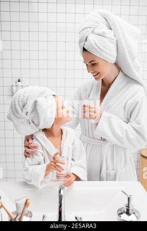 Lächelnde Mutter im Bademantel und Handtuch mit einem Glas Wasser In der Nähe Tochter im Bad Stockfoto