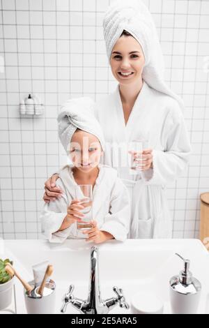 Lächelnde Mutter und Tochter in Bademänteln und Handtüchern mit Gläsern Von Wasser im Badezimmer Stockfoto