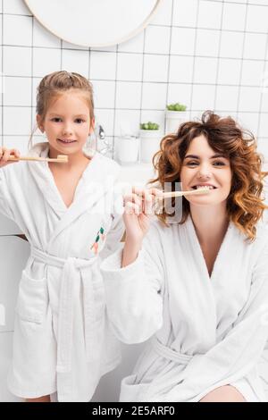 Mutter und lächelndes Kind in Bademänteln, die Zähne putzen und aussehen Bei der Kamera Stockfoto