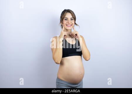 Junge Sportlerin schwanger trägt Sportkleidung auf weißem Hintergrund lächelnd zuversichtlich Zeigen und Zeigen mit den Fingern Zähne und Mund Stockfoto