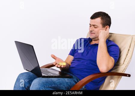 Ein älterer gehörloser Mann in einem Lederstuhl mit Laptop und einem Lächeln zeigt ein Hörgerät an der Kamera. Online Gesundheit Motivations Blog. Weißer Hintergrund. Stockfoto