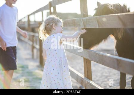 Kleines blondes Mädchen streichelte Pony im Zoo, ihr Bruder beobachtete sie und lächelte Stockfoto