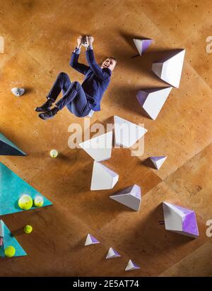 Junger Mann im Anzug klettert schwierige Route auf künstliche Wand in Boulderhalle. Konzept der beruflichen Herausforderungen. Der Geschäftsmann ist glücklich, dass er Reichweite hat Stockfoto