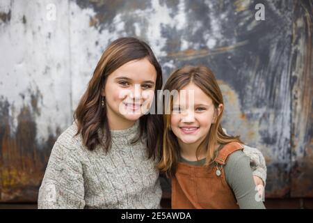 Zwei junge Schwestern sitzen nebeneinander und umarmen sich Mit den Armen umeinander im Winter sitzen In der Nähe einer städtischen Lagerhauswand Stockfoto