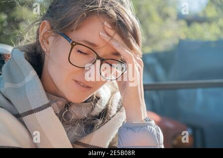 Kopfschmerzen, Migräne, Bluthochdruck. Junge Erwachsene Frau in einer Brille, die ihren Kopf hält, während sie im Freien sitzt, eingewickelt in ein Karli Stockfoto