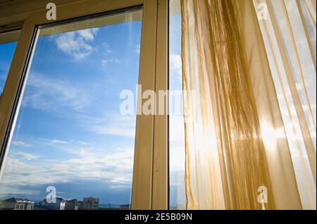 Die Sonne scheint durch Vorhänge und Fenster. Sonnendurchflutete Wohnung bei Sonnenuntergang. Blick auf die Stadt und blauer Himmel. Stockfoto