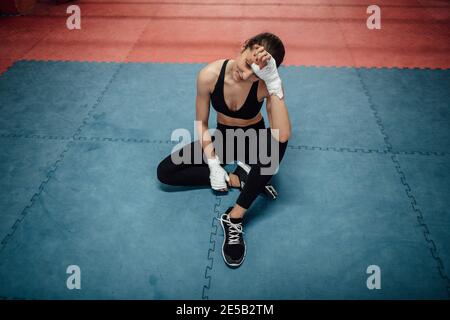 Ein Porträt einer jungen Sportlerin, die sich nach einem Workout im Fitnessstudio entspannt. Eine erschöpfte Sportlerin, die auf einer Matte sitzt und eine Pause vom Training macht. Stockfoto