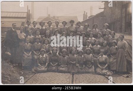 Große Gruppe von Arbeiterinnen aus der Zeit des Ersten Weltkriegs (und ein Mann), die außerhalb ihrer Fabrik fotografiert wurden (Straßenbahnen oder Eisenbahnschienen sind auf der Straße zu sehen). Die Frauen sind alle in Arbeitskleidung, die meisten tragen Kappen, um ihre Haare weg von der Maschine zu halten. Einige der Sitzenden (vor allem in der ersten Reihe) sind jung, vielleicht Teenager. Stockfoto