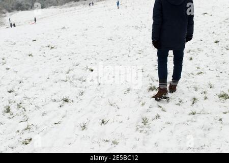 Nahaufnahme der beine und Füße von mans, die im Schnee stehen Auf einem Feldhang beobachten Menschen in der Ferne Rodeln Und im Winter draußen spielen Stockfoto