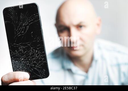 Telefon mit EINEM defekten Bildschirm Nahaufnahme. Ein weißer Mann hält ein schwarzes Smartphone mit einem geknackten Display. Stockfoto