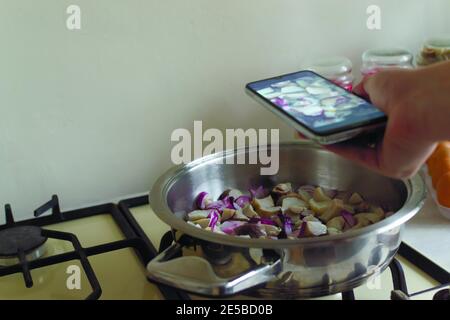 Eine Frau fotografiert Steinpilze und rote Zwiebeln in einer Pfanne in ihrer Küche. Food-Blogger. Social-Media-Konzept. Horizontale Ausrichtung. Kopieren Stockfoto