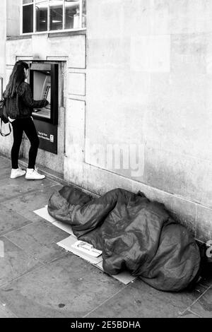 Eine obdachlose Person schläft in EINEM Schlafsack, während EINE junge Frau Geld an einem Geldautomaten in der Borough High Street, London, England abzieht. Stockfoto