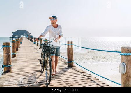 Porträt eines glücklichen lächelnden barfuss-Mannes, gekleidet in leichte Sommerkleidung und Sonnenbrille, der mit einem Fahrrad auf dem hölzernen Seebrücke läuft. Sorgloser Urlaub Stockfoto