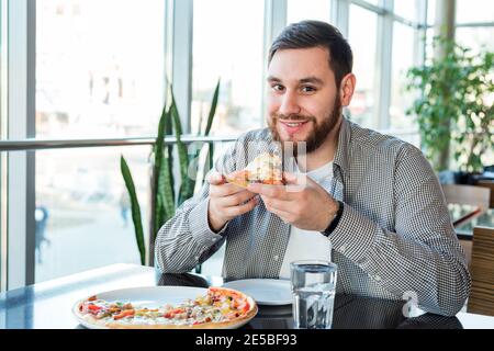 Glücklich lächelnd kaukasischen Mann essen italienische Pizza in der Pizzeria hält soziale Distanz. Leckere Pizza im Café. Stockfoto