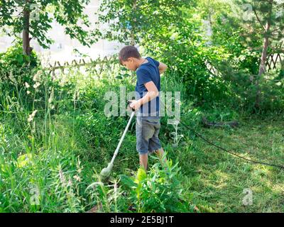 Junge, 11 Jahre alt, mäht den Rasen mit einer elektrischen Sense in der Nähe der Bäume im Hof eines Hauses an einem sonnigen Sommertag. Stockfoto
