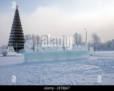 Die Inschrift auf Russisch - Frohes Neues Jahr! Aus Eis geschnitzt steht vor der Kulisse eines Weihnachtsbaums mit dem Wahrzeichen der Stadt. Stockfoto