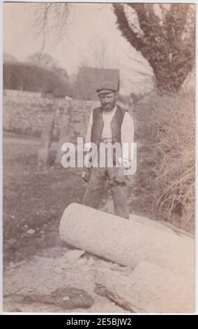 Steinmetz steht mit Hammer und Meißel in den Händen, neben einem runden Steinblock. Grabsteine sind im Hintergrund zu sehen, 1900er / 1910er Stockfoto