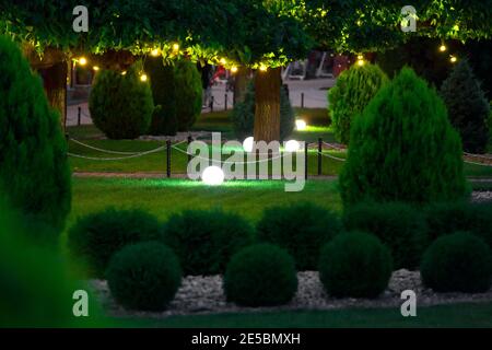 Beleuchtung Gartenleuchte mit elektrischer Erdlaterne mit Kugeldiffusor Lampe auf Rasen mit Girlande von warmen Glühbirnen an Baum Zweige mit Blättern l Stockfoto