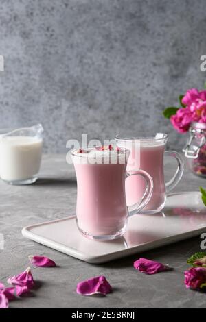 Rose Mond Milch in Glas Tassen und fallende Rosenblätter auf grauem Hintergrund. Nahaufnahme. Vertikales Format. Stockfoto