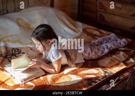 Ein Mädchen liegt auf dem Bauch auf einer Decke auf dem Bett und liest ein Buch und blättert durch die Seiten im Schlafzimmer eines Holzdorfhauses Stockfoto